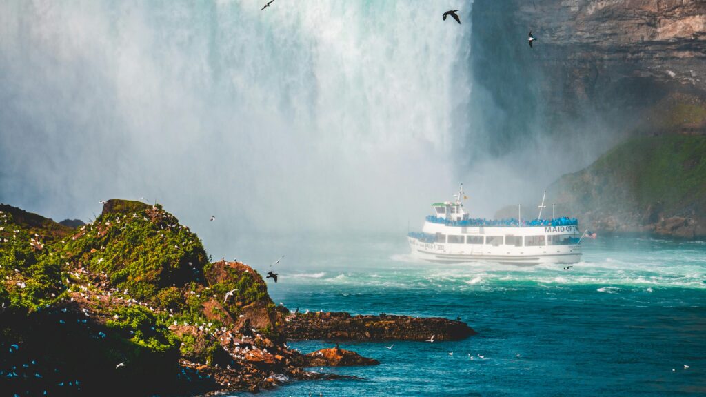 A scenic view of Niagara Falls featuring the Maid of the Mist ferry with lush greenery and birds.